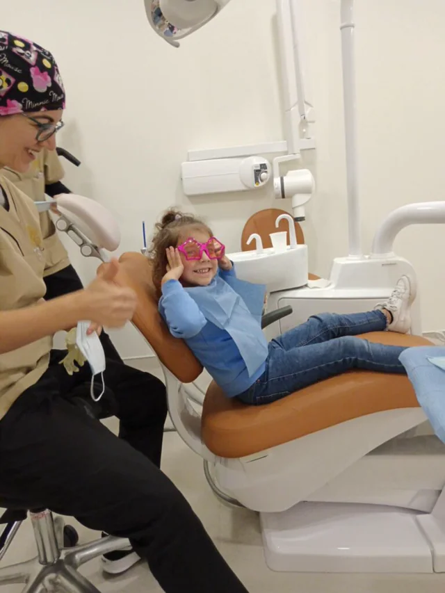 Criança com óculos coloridos na cadeira dentária, acompanhada por uma profissional